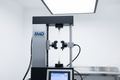 Calibrated tensile test frame with catheter shaft sample mounted between grips, test data on digital controller screen, M4D logo badge, white laboratory background